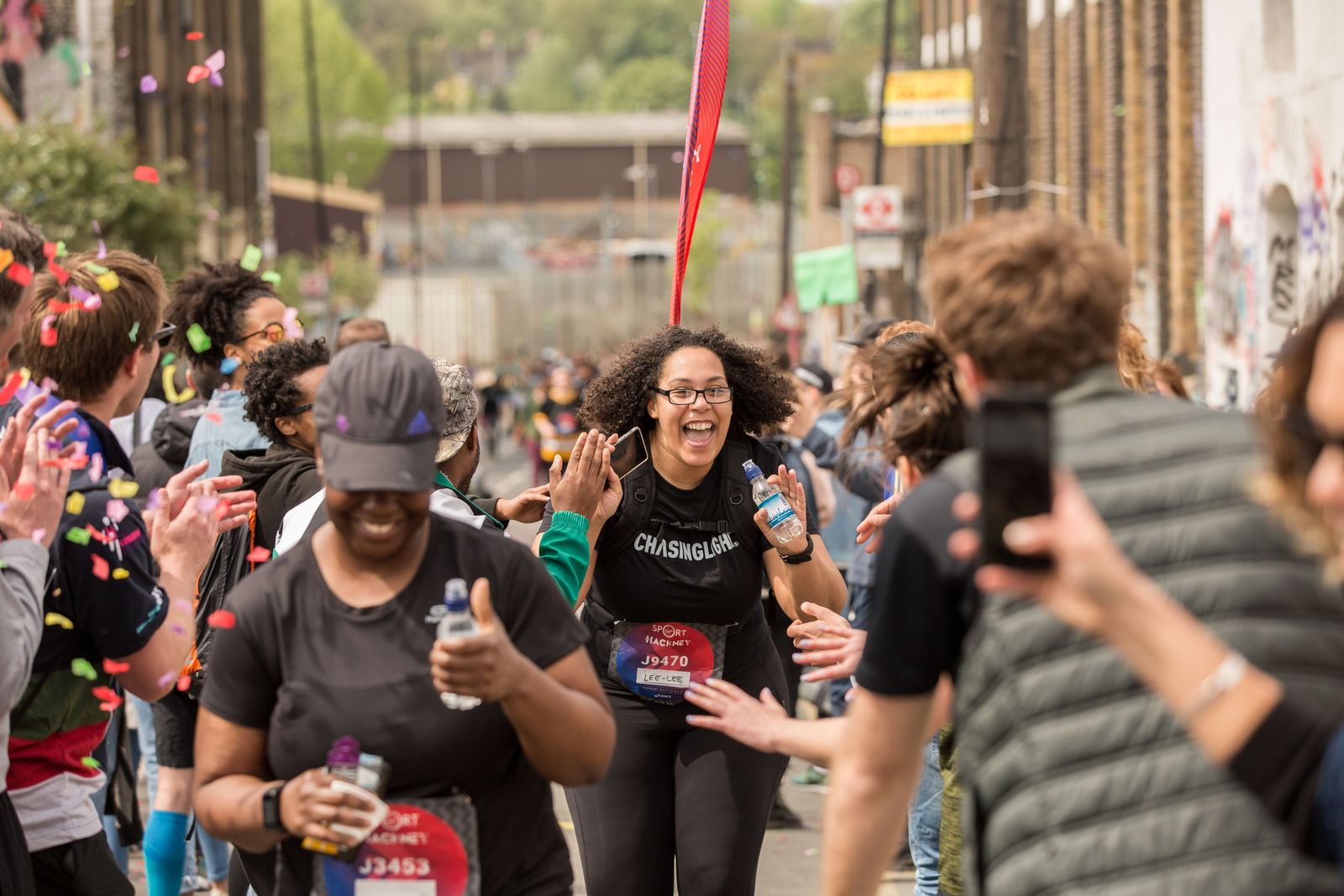 A Black British woman crosses the finish line at the Hackney Half Marathon in London in a shower of confetti while spectators cheer her on.